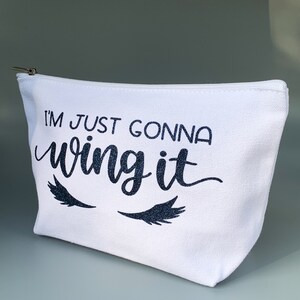 May include: White canvas makeup bag with a black zipper. The bag has the text "I'm just gonna wing it" in black glitter lettering with two black glitter eyelashes on either side of the text.