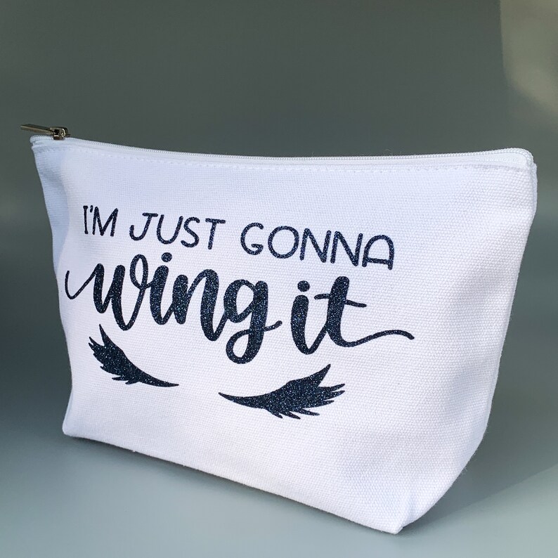 May include: White canvas makeup bag with a black zipper. The bag has the text "I'm just gonna wing it" in black glitter lettering with two black glitter eyelashes on either side of the text.