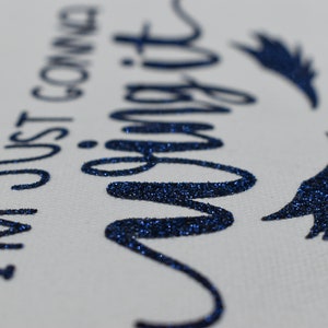 May include: A white canvas tote bag with the text "I'm just gonna wing it" in blue glitter lettering. The text is in a cursive font and there are two blue glitter wings on either side of the text.