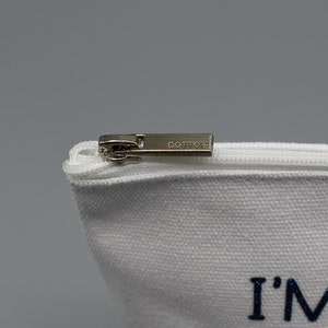 May include: White canvas zippered pouch with a silver zipper pull. The pouch has the text "I'M JUST" printed in blue glitter.
