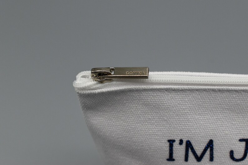 May include: White canvas zippered pouch with a silver zipper pull. The pouch has the text "I'M JUST" printed in blue glitter.