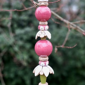Könnte beinhalten: Ein vertikaler, dekorativer Gartenstecker mit einer Reihe von rosa Keramikkugeln, weißen blütenförmigen Elementen und einer spitzen Spitze. Der Stecker ist auf einem Holzsockel montiert und schafft einen skurrilen Akzent im Freien. Ca. 100 cm hoch.