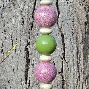 Könnte beinhalten: Ein skurriler Gartenstecker mit einer goldenen Krone auf einer grünen Keramikkugel. Der Stecker setzt sich mit abwechselnd rosa gemusterten und grünen Kugeln fort, die durch kleine, gesprenkelte Steine getrennt sind. Der Stecker ist auf einem Holzsockel montiert.