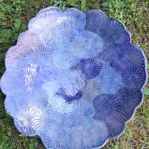 Könnte beinhalten: Eine runde Keramikschale mit gewelltem Rand und einer glänzenden Glasur in verschiedenen Blautönen. Die Oberfläche ist mit einem geprägten Muster aus zahlreichen gänseblümchenartigen Blumen verziert, was für eine strukturierte Optik sorgt.