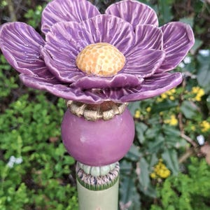 Könnte beinhalten: Handgefertigter Keramik-Gartenstecker mit einer großen, lila Blume mit strukturiertem, gelbem Zentrum. Der Stecker hat eine Reihe von lila und grünen Elementen, darunter ein Blattdesign. Ein dekoratives Gartenaccessoire.