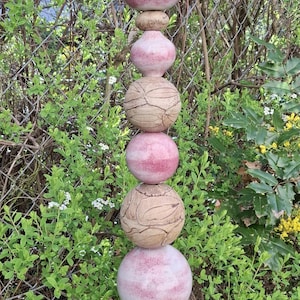 Könnte beinhalten: Ein dekorativer Gartenstecker mit gestapelten Keramikelementen. Die Spitze ist tropfenförmig, gefolgt von Kugeln und einem strukturierten Abschnitt. Der Stecker ist hauptsächlich rosa und beige, mit einer schwarzen Metallstange. Höhe ca. 150 cm.