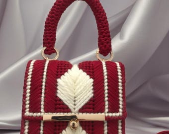 May include: A handcrafted red and white handbag with a braided handle and gold-tone hardware. The bag features a geometric design with a central white leaf-like pattern. The handle is approximately 6 inches long.