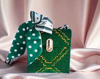 May include: A green woven handbag with gold accents and a gold clasp. A green and white polka dot scarf is tied to the top of the bag. The bag is sitting on a pink satin surface.