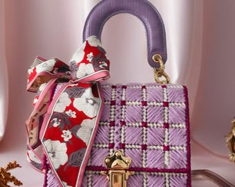 May include: A lavender handbag with a stitched grid pattern, a purple handle, and a gold clasp. A floral scarf in red, white, and pink is tied to the handle. The bag has a structured shape and a decorative design.