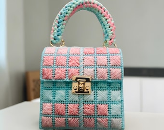 May include: A small, square handbag with a grid pattern in shades of pink and turquoise. The handle is a curved arch in matching colors. The bag features a gold-colored clasp and hardware. The overall design is handmade and unique.