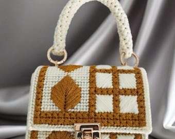 May include: A handcrafted white and gold handbag with a woven design. The bag features a cream-colored handle and gold-toned hardware, including a clasp and ring details. The front of the bag has a leaf design.