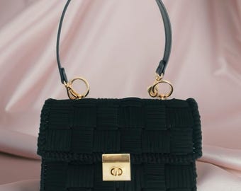May include: A black handbag with a woven design. The bag features a gold-colored clasp and hardware, with a black handle. The bag is rectangular in shape and has a structured design. The background is a soft pink.
