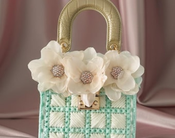 May include: A mint green and white checkered handbag featuring a structured gold-toned handle with a textured finish. The front is adorned with three large, layered cream-colored fabric flowers, each centered with a cluster of small white faux pearls.