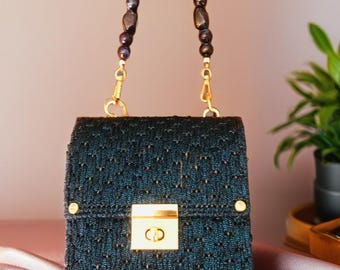 May include: A small, square, dark teal handbag with a beaded handle. The bag has a gold clasp and gold hardware. The handle is made of dark, irregularly shaped beads. The bag is sitting on a pink satin surface.