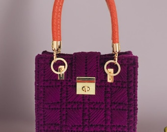 May include: A structured, purple handbag with a quilted pattern. It features a braided orange handle and gold-colored hardware, including a clasp closure. The bag's design is boxy and elegant.
