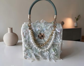May include: A unique handbag with a textured, light blue and white exterior. It features a gold chain and pearl strap, a gold rose clasp, and a green handle. The bag is sitting on a white surface.