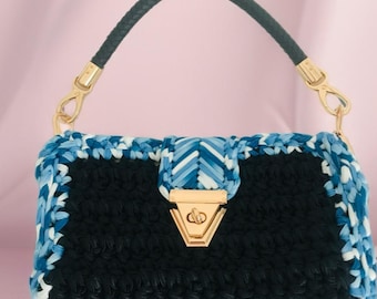 May include: A black crochet handbag with a braided blue and white trim and a black leather handle. The bag features a gold-colored clasp and hardware. The bag is approximately 10 inches wide.