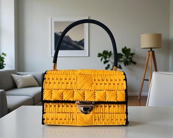 May include: A yellow handbag with a woven design, black trim, and a black handle. The bag features a silver clasp and gold-colored accents. The bag is sitting on a white table.
