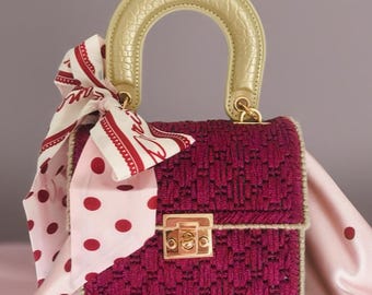 May include: A structured, magenta handbag with a gold-toned handle and clasp. A patterned scarf in white and red polka dots is tied to the handle. The bag has a woven texture and a rectangular shape, suggesting a fashion accessory.