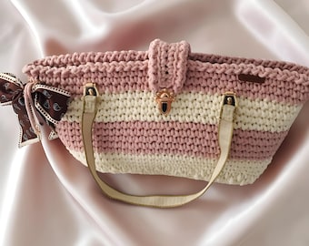 May include: A handmade crochet tote bag with alternating stripes of light pink and cream. The bag features a flap closure with a gold-toned clasp, cream-colored straps, and a decorative brown ribbon tied to the side. The bag is designed for carrying items.