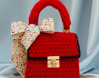 May include: A handmade red crochet handbag with a matching handle and gold-colored clasp. A floral patterned bow is tied to the side of the bag. The bag has a black trim.