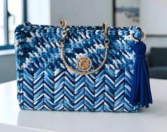 May include: A blue and white woven handbag with a gold chain handle and a gold rose embellishment. The bag has a blue tassel and gold hardware. The bag is rectangular and sits on a white surface.