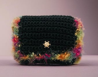May include: A small, black crochet bag with a textured surface. The edges are trimmed with colorful, fluffy yarn in shades of pink, yellow, and green. A gold-colored button with a pearl-like center secures the flap.