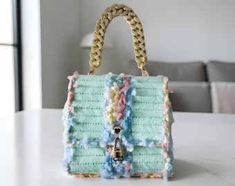 May include: A pastel green handbag with a gold chain handle. The bag features a textured, woven design with colorful, fluffy accents in pink, blue, and yellow. The clasp is gold-toned.