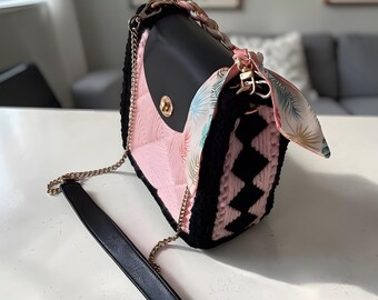 May include: A black and pink handbag with a gold chain strap and a decorative scarf. The bag features a black flap with a gold clasp, pink quilted interior, and black crocheted sides with a geometric pattern. The scarf has a tropical leaf print.