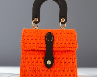May include: A bright orange crochet handbag featuring a structured rectangular body with a woven texture. It includes a solid black arched handle attached with gold-toned metal rings and a black leather-like tab closure with a gold-toned snap button.