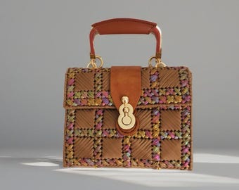 May include: A structured handbag featuring a woven brown leather body with a grid pattern interspersed with metallic pink, gold, and green threads. It includes a solid brown leather top handle, a matching brown leather flap closure, and gold hardware.