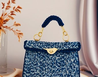 May include: A structured handbag crafted from blue and white variegated yarn with a crochet texture. The bag features a solid dark blue crochet handle, gold-toned metal hardware, and a distinctive gold bird-shaped clasp on the front flap.