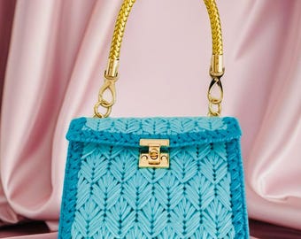 May include: A structured handbag featuring a light blue crocheted body with a darker blue border and a gold-toned metal clasp. The bag includes a single braided gold-toned handle attached with gold-toned hardware, creating a distinct contrast.