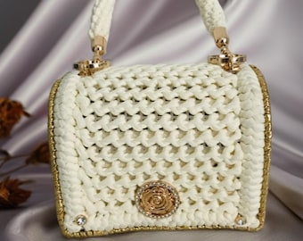 May include: Cream-colored, hand-crocheted handbag with a woven pattern. Features a gold-toned frame and accents, including a rose-shaped embellishment and small jewels. The handle is also cream-colored and crocheted.