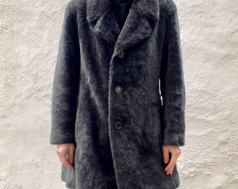 May include: A dark gray, mid-length coat with a textured, fuzzy appearance. The coat features a wide collar, three buttons, and a tailored fit. The person is wearing matching gray pants. The coat appears to be made of a soft, warm material.