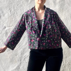 Op de afbeelding: Een getailleerd jasje met bloemenprint, knoopsluiting en lange mouwen. Het jasje is zwart met roze en groene bloemen.