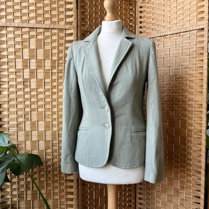 Puede incluir: Una chaqueta verde claro con un cierre de botón único y dos bolsillos. La chaqueta está en un maniquí contra un fondo tejido.