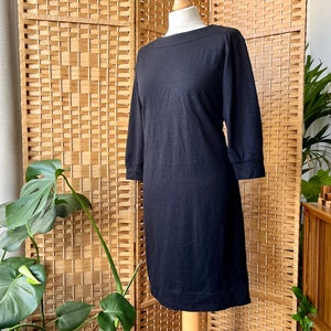 May include: A black, knee-length dress with three-quarter sleeves. The dress has a boat neckline and a simple, classic design. The dress is displayed on a mannequin against a natural wood background, with green plants in the foreground.