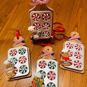 May include: Wooden cutting boards with metal candy molds, each holding peppermint candies in red, white, and green. Gingerbread man ornaments and red and white striped bows add a festive touch. Perfect for Christmas decor.