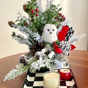 May include: A festive centerpiece featuring a white owl, pinecones, red berries, and greenery. The arrangement sits on a black and white checkered tray with two candles. A red and black ribbon adds a decorative touch.