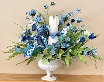 Peut inclure: Un arrangement floral de Pâques avec un lapin en céramique blanche orné d'un collier floral bleu. Il comprend des fleurs bleues et blanches, de la verdure et un ruban à carreaux, le tout dans une urne blanche vieillie. Décoration de Pâques.