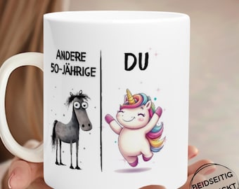 May include: White ceramic mug with a whimsical design. The mug features a split design with a sad horse on the left and a happy unicorn on the right. Text includes "ANDERE 50-JÄHRIGE" and "DU". The mug is held by a person.