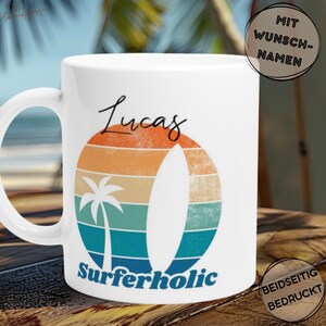 Op de afbeelding: Witte keramische mok met een retro zonsondergangontwerp, een surfboard en een palmboom. De mok heeft de naam "Lucas" in script boven het ontwerp en het woord "Surferholic" eronder. De mok heeft een wit handvat.