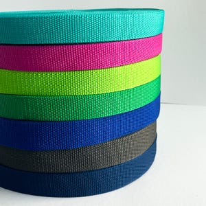 Könnte beinhalten: Ein Stapel farbenfroher Nylon-Gurtbandrollen in Türkis, Pink, Limettengrün, Grün, Blau, Grau und Marineblau. Die Rollen sind eng gewickelt und zeigen die gewebte Textur des Materials. Der Hintergrund ist schlicht weiß.