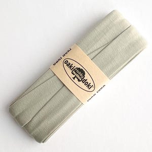 May include: A folded length of sage green bias tape wrapped in a tan paper band. The band features the black text Oaki Doki Hobby Textiles with a tree logo. The fabric has a visible woven texture and is neatly coiled into a flat, rectangular shape.