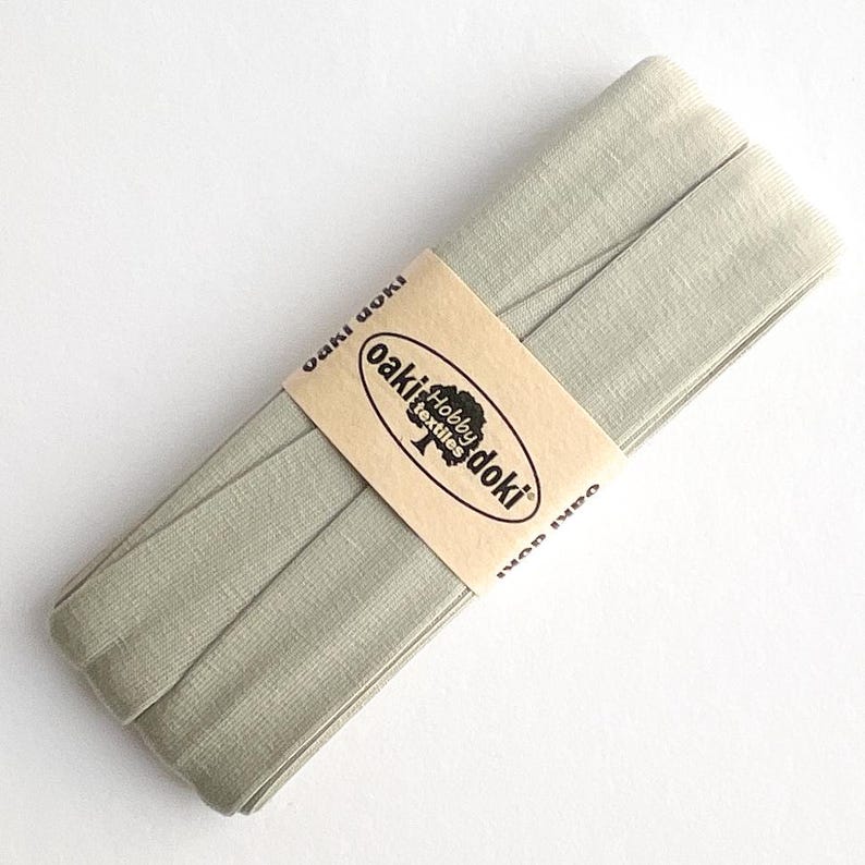 May include: A folded length of sage green bias tape wrapped in a tan paper band. The band features the black text Oaki Doki Hobby Textiles with a tree logo. The fabric has a visible woven texture and is neatly coiled into a flat, rectangular shape.
