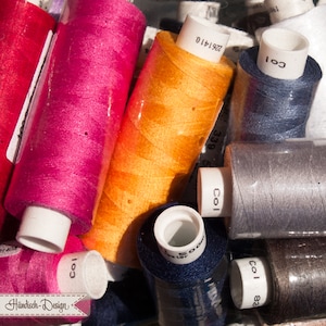 Könnte beinhalten: Nahaufnahme einer Vielzahl von Garnrollen in verschiedenen Farben, darunter Pink, Orange, Blau, Grau und Braun. Die Spulen sind mit Zahlen und dem Wort "Col." beschriftet.