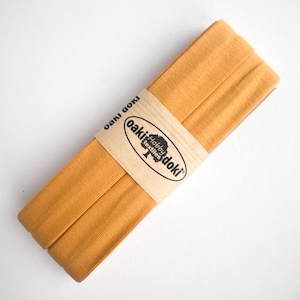 May include: A folded strip of mustard yellow fabric tape secured with a tan paper band. The band features the black printed text "oaki doki" and "Hobby textiles" with a tree icon. The fabric has a visible ribbed texture and is neatly layered.