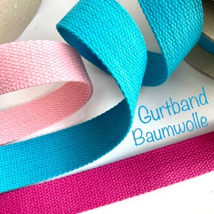 May include: Four rolls of cotton webbing in pink, turquoise, and fuchsia. The webbing is woven and has a textured surface. The text "Gurtband Baumwolle" is visible in the image.