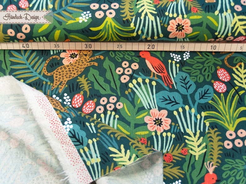 May include: A dark green fabric featuring a repeating pattern of orange leopards, red birds, pink flowers, and various green foliage. A wooden ruler is placed across the top, showing measurements from 5 to 40 centimeters. The fabric has a white reverse side.